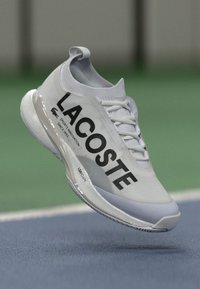 Fehér sportcipő „LACOSTE” logóval, sima felső anyaggal, gumitalppal és textúrázott részletekkel, sporttevékenységekhez tervezve.