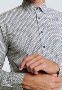 Weißes Hemd mit knöpfen und Punktkragen, das ein blaues geometrisches Muster zeigt. Die langen Ärmel und die strukturierten Stoffdetails verleihen dem Shirt einen besonderen Charme.