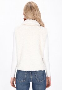 Vrouw met lange blonde haren die een witte textuurloze mouwloze vest over een langemouwen shirt en blauwe spijkerbroek draagt, met de rug naar de camera gekeerd.