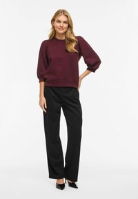 Femme aux longs cheveux blonds portant un haut bordeaux à manches bouffantes, un pantalon noir et des chaussures noires à talons, debout devant un fond blanc.