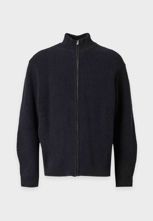 Jeune homme debout portant un cardigan noir zippé, un t-shirt blanc, un pantalon ample foncé et des baskets noires sur un fond clair uni.