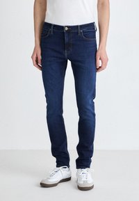 Jeans skinny bleu foncé en denim, avec une fermeture à bouton à l'avant, cinq poches et des détails de couture jaune contrastant.