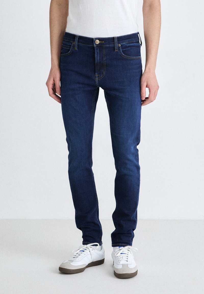 Jeans skinny bleu foncé en denim, avec une fermeture à bouton à l'avant, cinq poches et des détails de couture jaune contrastant.