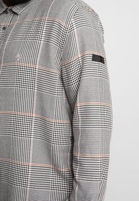 Camisa de manga larga con un patrón de pata de gallo en blanco y negro, con líneas naranjas, un collar y una pequeña etiqueta negra en la manga.