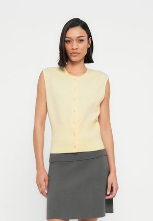 Lys gul ermeløs cardigan med rund hals, med oransje knapper og ribbet kant, sammen med et mørkegrått skjørt.