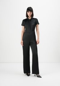 Woman standing with straight posture wearing a black short-sleeve jumpsuit and black pointed high heels on a white background.