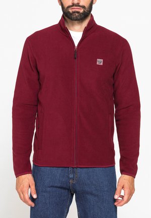 Uomo che indossa una giacca in pile bordeaux con cerniera e un piccolo logo rettangolare sul lato sinistro del petto, abbinata a jeans blu.