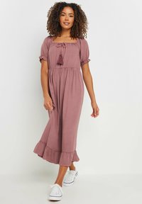 Vestido midi rosa con escote cuadrado, mangas abullonadas, cordón con borlas y dobladillo con volantes; combinado con zapatillas blancas.