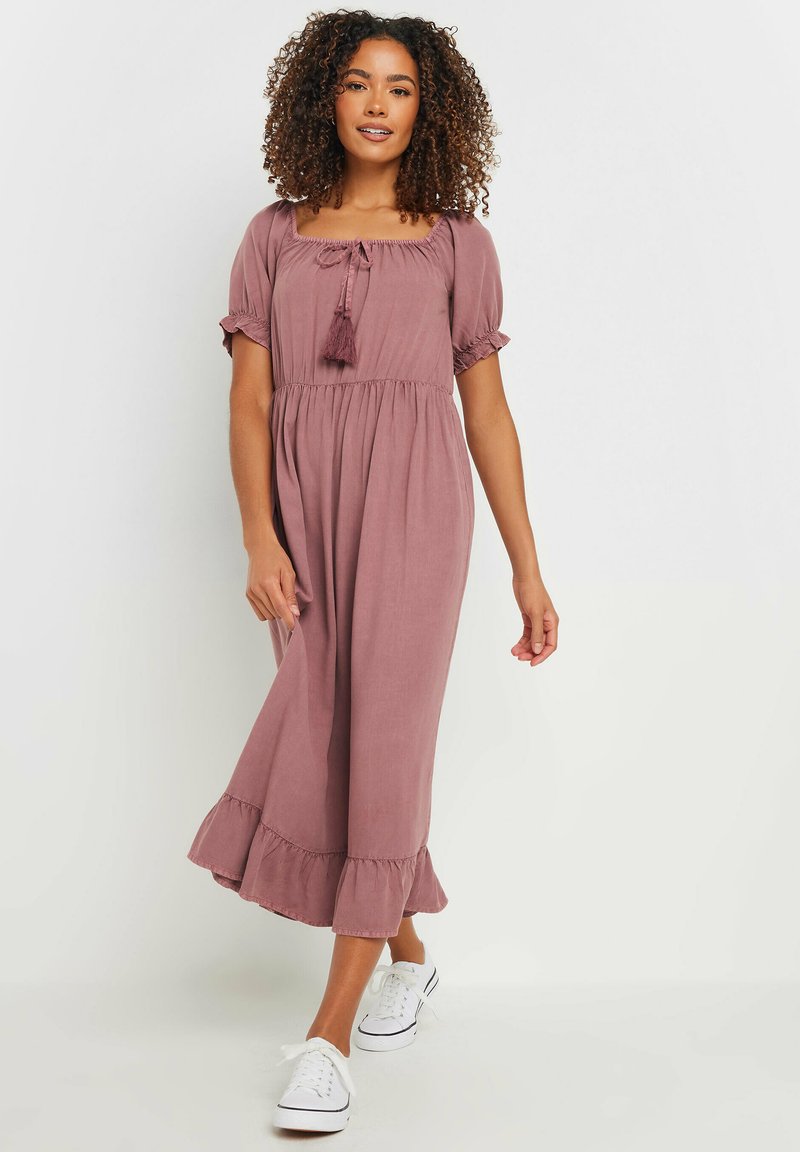 Vestido midi rosa con escote cuadrado, mangas abullonadas, cordón con borlas y dobladillo con volantes; combinado con zapatillas blancas.