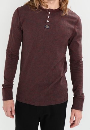 Langærmet burgunder henley-skjorte lavet af blødt stof, med en knappet halsudskæring og et lille logo-mærke nær kraven.