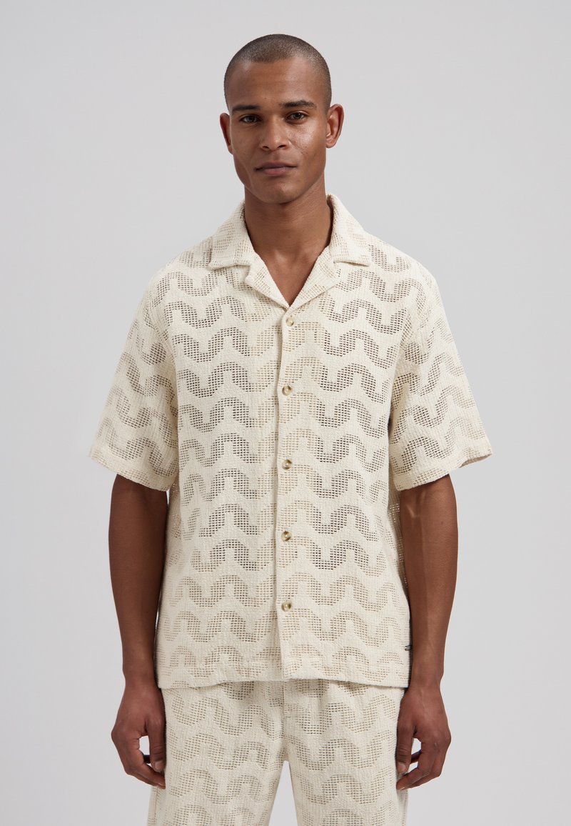Homme portant une chemise boutonnée à manches courtes couleur crème et un short assorti avec un motif en maille ondulée, debout devant un fond uni.