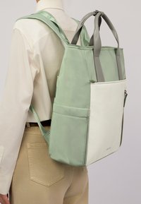 Zaino di colore verde chiaro e bianco, con doppie maniglie superiori, spallacci regolabili, texture liscia e una tasca frontale con zip.