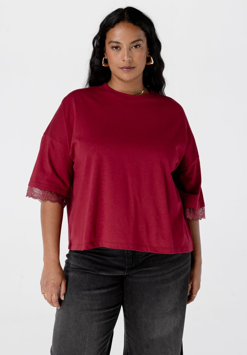 Vrouw met donker golvend haar, gekleed in een losse rode top met kantafgewerkte mouwen en zwarte jeans, staande tegen een effen witte achtergrond.