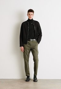 Chaqueta negra con cremallera sobre un cuello alto negro, combinada con pantalones cargo verde olivo y botas negras, con un corte slim y bolsillos prácticos.