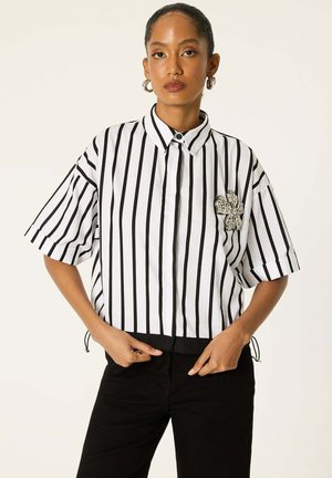 Femme portant une chemise à manches courtes à rayures verticales noires et blanches avec une broche florale ornée de bijoux sur la poitrine, associée à un pantalon noir.