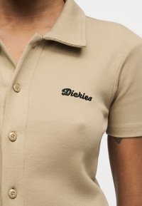 Bež polo majica s kratkimi rokavi z gumbi in črnim vezenim logotipom "Dickies" na levi prsih.