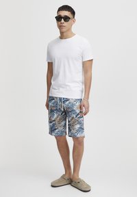 T-shirt blanc à manches courtes, short de bain bleu et beige à motifs avec des feuilles de palmier et des fleurs, chaussures gris à enfiler, et lunettes de soleil noires.