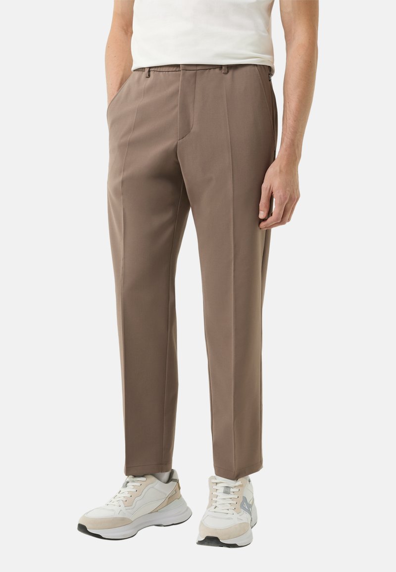 Bruine op maat gemaakte broek met een gladde textuur, voorzien van een platte voorkant en zijzakken, gecombineerd met witte en grijze sportschoenen.