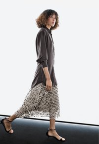 Vrouw met krullend haar die loopt, gekleed in een donkerbruine lange mouwen top, een transparante midi rok met pailletten en zwarte sandalen met lage hak.