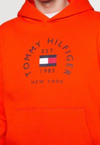 Oranje hoodie met een voorzak en een cirkelvormig logo met de tekst "TOMMY HILFIGER", oprichtingsdatum en een kleurblokvlagontwerp.