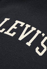 Crna tkanina s teksturiranom površinom s velikim krem bojom slovima koja ističu "LEVI'S" u središtu.