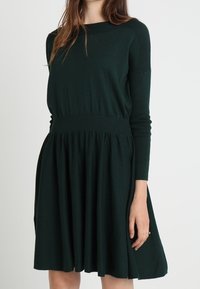 Mörkgrön stickad klänning med en figurnära bodice, ribbade ärmar och en svängd kjol. Materialet verkar mjukt och har en annan slät struktur.