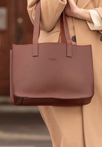 Brązowa skórzana torba na ramię o gładkiej fakturze, prostokątnym kształcie i podwójnych ramiączkach. Na przodzie znajduje się subtelne logo.