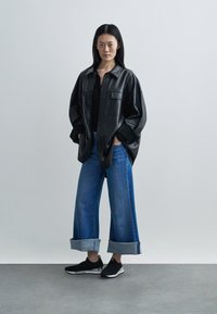 Camisa casaco de pele preta com um corte descontraído, combinada com jeans azuis de perna larga com bainhas enroladas, e ténis pretos sem atacadores.