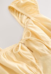 Camicia in tessuto leggero e giallo, con spalle arricciate, bordo in pizzo e fiocco davanti, caratterizzata da una finitura morbida e testurizzata.
