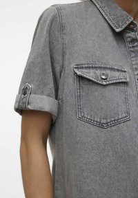 Primer plano de una persona que lleva una camiseta de mezclilla gris de manga corta con un bolsillo en el pecho abotonado y las mangas arremangadas.