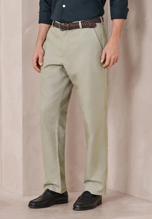 Mand iført lyse beige chinos, en mørk skjorte stukket ind, et fletbånd i brun samt sorte loafers, stående mod en ensfarvet baggrund.