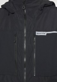 Veste noire avec fermeture éclair à l'avant, deux poches zippées, une capuche, et une étiquette indiquant "BURTON" avec un accent gris. Tissu lisse et texturé.