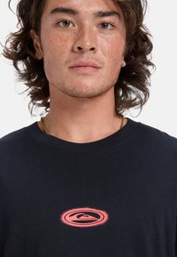 Dunkelblaues T-Shirt mit einem runden, verblassten rosa Logo auf der Brust. Der Stoff wirkt weich und hat einen klassischen Rundhalsausschnitt sowie eine lockere Passform.