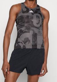 Top deportivo en gris oscuro con un patrón gráfico. Presenta un escote redondo, diseño sin mangas y un corte ajustado. Combinado con pantalones cortos negros.