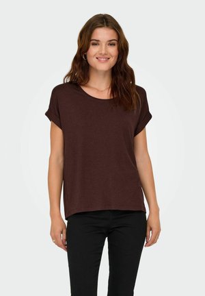 Giovane donna con capelli castani ondulati che indossa una maglietta marrone a maniche corte e pantaloni neri, sorridente con le mani lungo i fianchi.