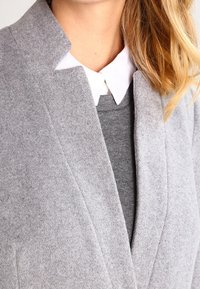 Chaqueta gris de mezcla de lana con cuello y solapa con muesca. Debajo, se ve un suéter gris acompañado de un cuello de camisa blanca.