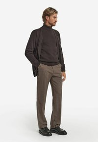 Dunkler Strickpullover mit Rollkragen unter einer locker sitzenden dunklen Strickjacke, kombiniert mit hellbraunen Karohosen und schwarzen Schnürschuhen.