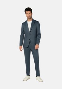 Dunkelblauer Anzug mit strukturierter Oberfläche, bestehend aus einem einreihigen Sakko, schmal geschnittenen Hosen und weißen Turnschuhen. Darunter ein weißes T-Shirt.