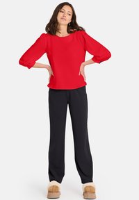 Blusa rossa con maniche tre quarti, scollatura rotonda e vestibilità rilassata, abbinata a pantaloni neri e scarpe beige con bordo di pelliccia.