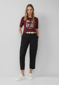 T-shirt graphique bordeaux à manches courtes, pantalon noir court, ceinture tissée crème avec boucle marron, et sneakers blanches.