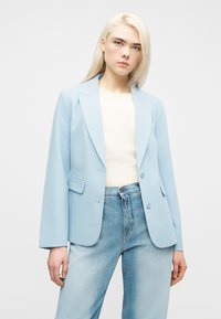 Νεαρή γυναίκα με ίσια ξανθά μαλλιά φοράει ανοιχτό μπλε σακάκι πάνω από μπεζ μπλούζα και ξεθωριασμένο μπλε τζιν, στέκεται μπροστά σε μονόχρωμο φόντο.