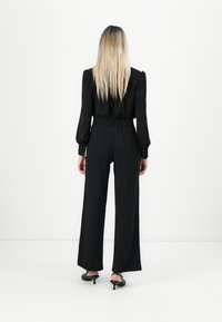 Femme aux longs cheveux blonds portant un blouse noire, un pantalon large à taille haute et des chaussures à talons noirs, se tenant contre un fond blanc, de dos.