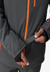 Graue Outdoor-Jacke mit orangefarbenem Reißverschluss und Akzenten. Mit Klettverschluss zur Anpassung der Ärmelbündchen. Nahaufnahme von Händen, die den Ärmel anpassen. Glatte Textur.