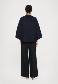 Cape en laine bleu marine avec un col rond, un design fluide et des manches amples, porté sur un pantalon large noir et des chaussures à talons.