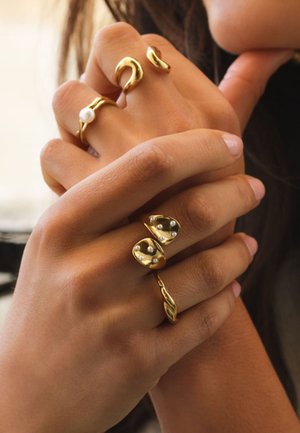 Manos entrelazadas cerca del rostro, con cuatro anillos de oro distintos con detalles de perlas, sobre un fondo neutro desenfocado.