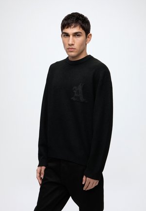 Jeune homme aux cheveux courts et foncés, portant un pull noir avec un discret motif en forme de lettre A sur la poitrine, debout devant un fond blanc uni.