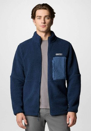 Giacca in pile blu navy con colletto alto, dotata di una tasca a toppa in blu chiaro con texture. Tessuto morbido con vestibilità rilassata.