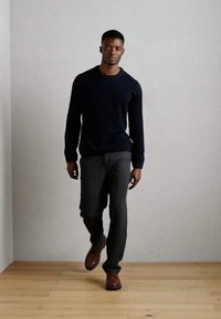 Navy knit sweater, gray tailored trousers, and brown shoes. Model walking on light wooden floor against a plain light wall.