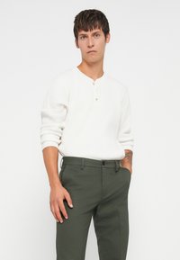 White, ribbed long-sleeve henley shirt with three buttons, paired with olive green tailored pants. Model standing with hands in pockets.