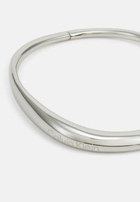 Silverfärgad metallhalsband med en slät, böjd design. Har graverad "Calvin Klein"-logotyp på ena sidan.